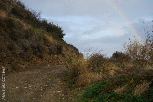 A rugged dirt path winding through the wild hills of Crete, featuring a vibrant rainbow stretching across the overcast sky.