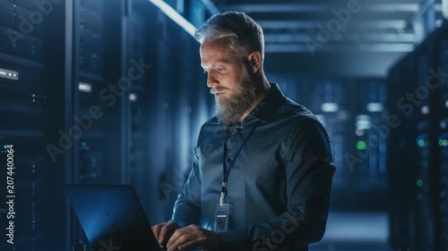 Middle-aged bearded man working on a laptop in a data center, illuminated by server rack lights, IT administrator managing server infrastructure, cybersecurity.