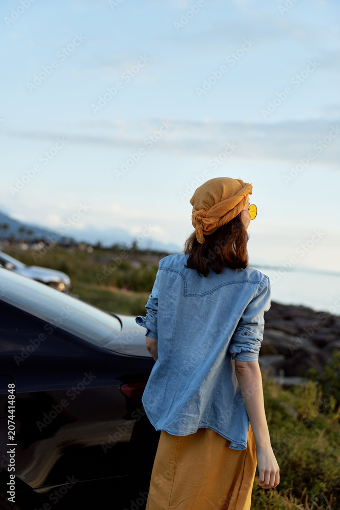 Fototapeta premium Young woman in a denim shirt and hat stands beside a parked car by the coast, capturing a casual outdoor lifestyle moment with soft daylight and distant sea.