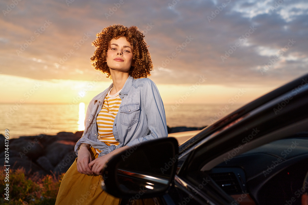 Fototapeta premium A woman with curly hair wearing a denim jacket and striped top stands beside a car at sunset, showcasing a relaxed, outdoor lifestyle moment suitable for travel and leisure themes.