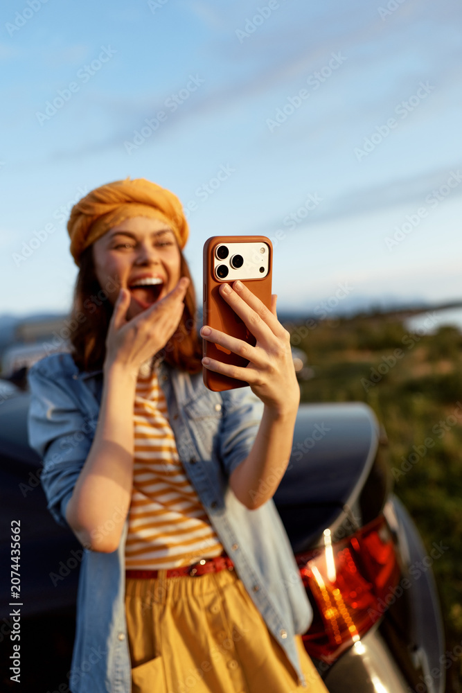 Fototapeta premium Cheerful woman outdoors taking a selfie, smiling with eyes opened, striped shirt, denim jacket, casual fashion, warm sunlight, vibrant colors, natural landscape, car in background, candid lifestyle