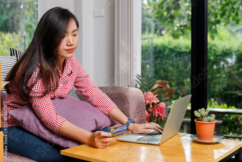 Young Asian woman holding credit card and using laptop for online shopping at home. Beautiful female entrepreneur paying with credit card on laptop computer in living room.