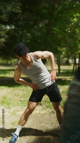 A young man wearing athletic attire performs warm-up stretches in a park. He is actively preparing his body for a workout amidst green trees and grass.