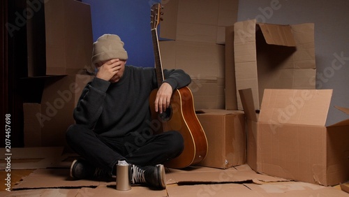 Canvas-taulu Young male musician sitting on cardboard boxes in a makeshift shelter with his a