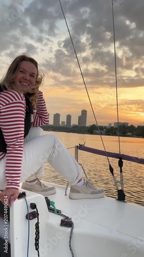 Smiling young woman enjoys sailing on recreational yacht at golden sunset, relaxing on deck with calm water. Yachting lifestyle, summer adventure, freedom, leisure travel, outdoor recreation