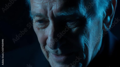 Elderly Caucasian man with gray hair smiles warmly, transitioning to a thoughtful expression, illuminated by soft blue lighting in a darkened setting