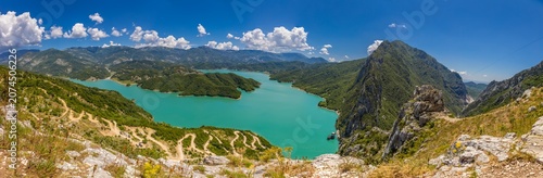 Wide-screen high-resolution panoramic view of Bovilla Lake in Albania, showcasing the striking turquoise waters surrounded by steep rocky cliffs and rugged mountain terrain. The reservoir  Bovilla