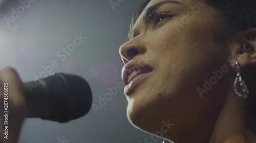 Female singer with dark hair passionately performs into a microphone under bright stage lights, showcasing her expressive emotions and engagement with the audience in a music venue