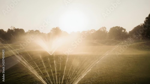 Watering system sprays mist over lush green golf course grass at sunrise, creating a serene atmosphere with sunlight filtering through trees in the background