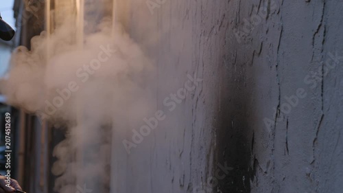 Close up of person's hand exhaling thick white vapor, illustrating atmospheric release and transient urban mystery against a textured wall.
