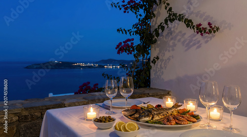 Romantic seaside dinner table at sunset with candlelight, seafood platter, and wine glasses overlooking the calm Mediterranean coast under a deep blue evening sky.