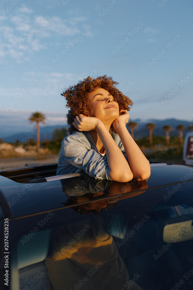 Fototapeta premium A woman enjoys a sunny day, leaning from car windows with a relaxed smile, embodying casual lifestyle and outdoor freedom, capturing a carefree travel vibe against a blue sky.