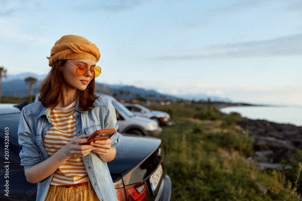 Fototapeta premium A stylish woman beside a car on a coastal road checks her phone, wearing a denim jacket, striped shirt, orange hat and yellow sunglasses. Concept for travel, leisure, and lifestyle.