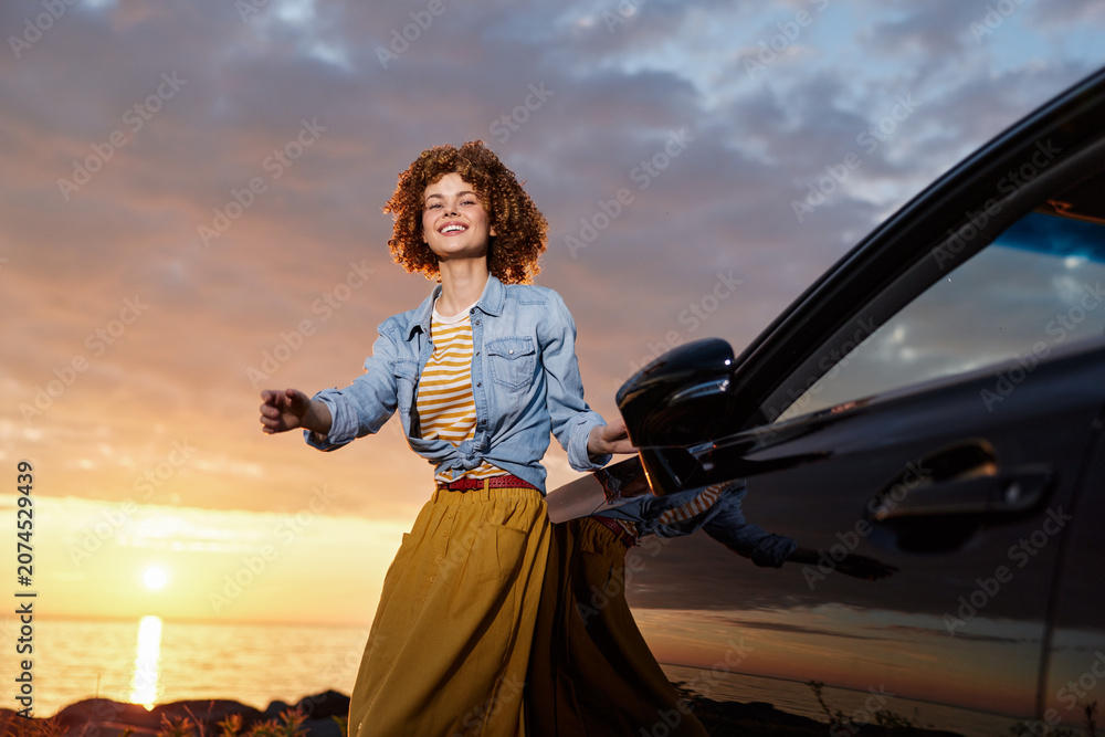 Fototapeta premium Joyful woman in denim jacket and yellow skirt poses beside a car at sunset on a coastal beach, embracing a carefree lifestyle, breeze, and warm golden light