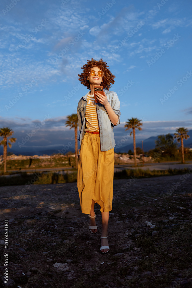 Fototapeta premium Cheerful woman outdoors walking on rocky ground, sunlit mood, casual summer dress, light jacket, palm trees in background, natural colors, relaxed pose, golden hour glow