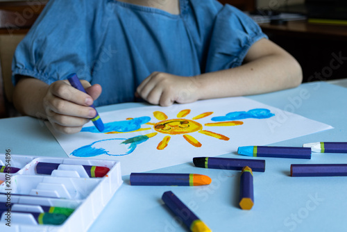 Child in blue shirt creating sun artwork with blue oil pastel, white paper with yellow sun and clouds, organized supplies in white box, scattered crayons on table.