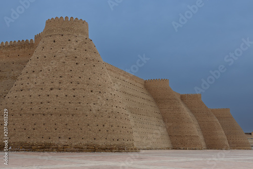 Ark Fortress walls in Bukhara with massive sloping buttresses and crenellated parapets under an overcast sky. This iconic Uzbekistan landmark image is ideal for travel publications and heritage.