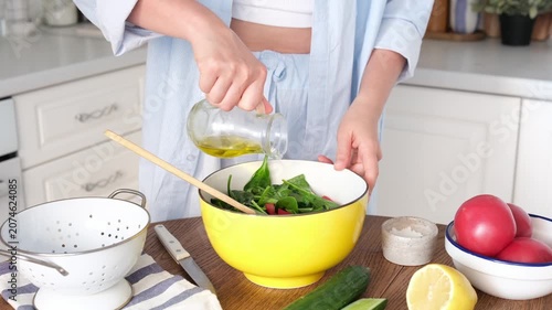 Woman in a light blue shirt pours olive oil into a yellow bowl with greens. Fresh vegetables, lemon, and kitchen utensils on a wooden table. Healthy eating concept. High quality photo