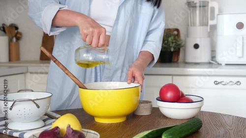 Woman in a light blue shirt pours olive oil into a yellow bowl with greens. Fresh vegetables, lemon, and kitchen utensils on a wooden table. Healthy eating concept. High quality photo