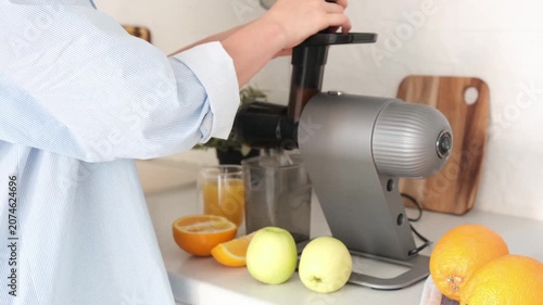 A person uses a countertop juicer to prepare fresh orange juice beside whole fruit, cut citrus pieces, a glass, and a small plant in a bright home kitchen.