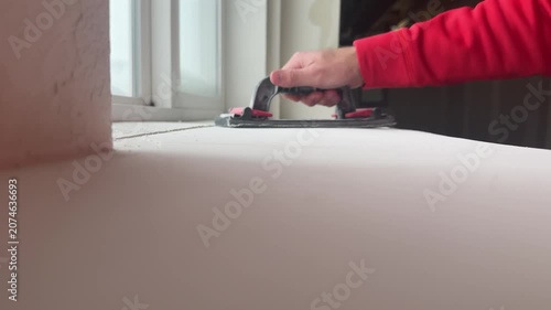 Worker sanding drywall compound with a hand sanding tool to create a smooth wall surface during interior renovation, producing fine dust in a home construction project.
