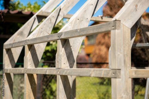 A timber greenhouse frame stands outdoors in bright daylight. Triangular rafters and cross bracing are pre drilled and screwed, with saw marks and joinery details visible.