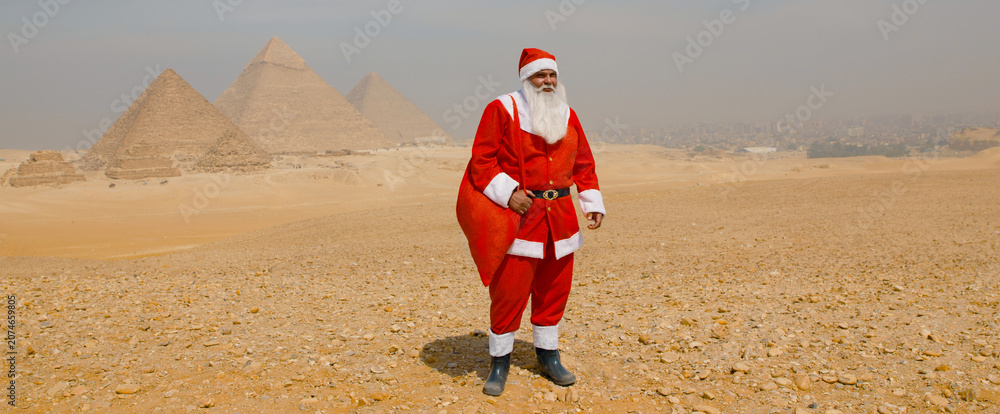 Fototapeta premium Whimsical Santa Claus in red costume stands holding a gift sack before the Giza pyramids in the sunny Egyptian desert ideal for cross cultural holiday