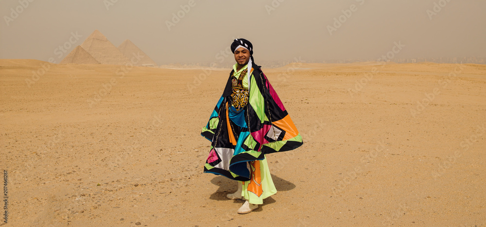 Fototapeta premium Joyful male Egyptian folk dancer in a bright multicolored tanoura costume with flowing cape standing in the desert before the Giza pyramids ideal for
