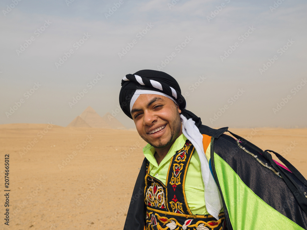 Fototapeta premium Cheerful Egyptian folk dancer in colorful embroidered tanoura costume and black turban smiling warmly in the desert near the Giza pyramids ideal for
