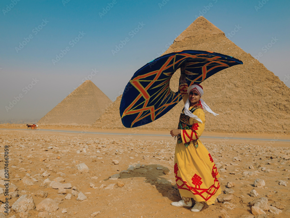 Fototapeta premium Joyful Egyptian tanoura folk dancer twirling a colorful skirt overhead before the Giza pyramids in the sunlit desert ideal for Egypt tourism culture