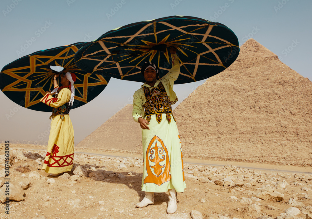 Fototapeta premium Vibrant Egyptian tanoura dancers lift and spin colorful embroidered skirts in a festive folk performance before the Giza pyramids ideal for MENA