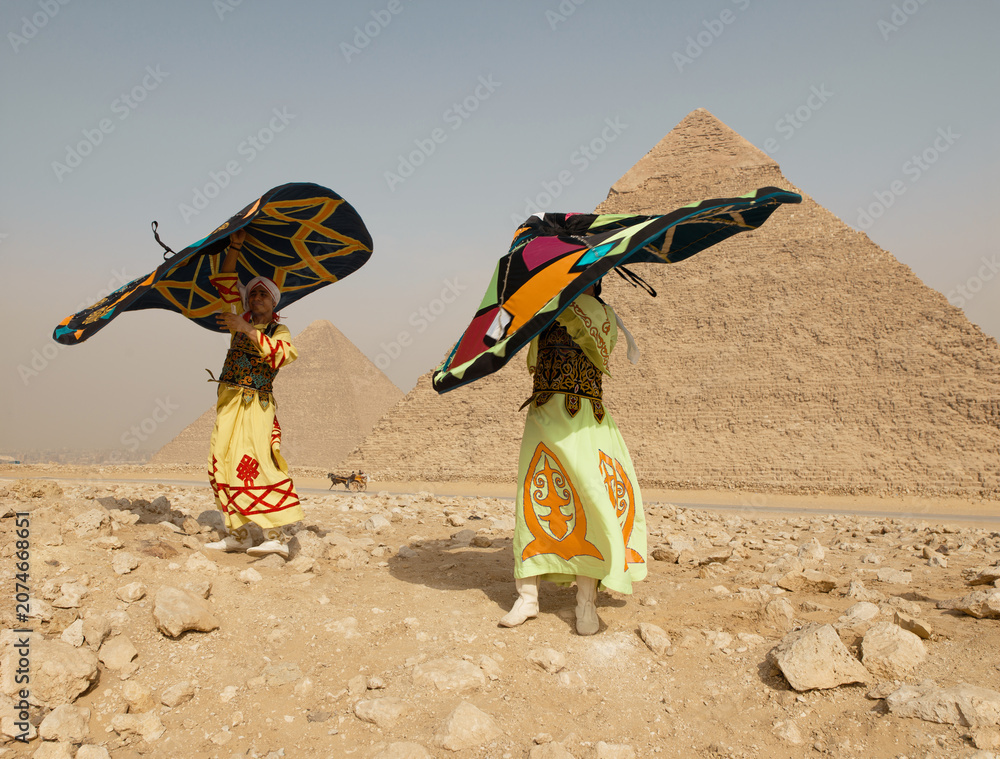Fototapeta premium Vibrant Egyptian tanoura folk dancers spinning brightly colored skirts overhead in a festive sufi performance before the Great Pyramids of Giza in the