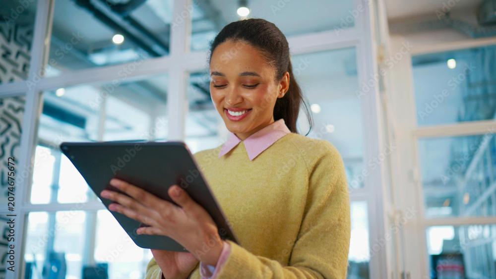Fototapeta premium Successful business woman holding tablet and smiling while reading message. Standing near glass office wall. Female checking online project updates. Corporate technology lifestyle concept.