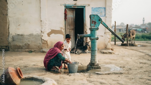 Woman draws clean water from a hand pump while a young child watches in a rural village setting.