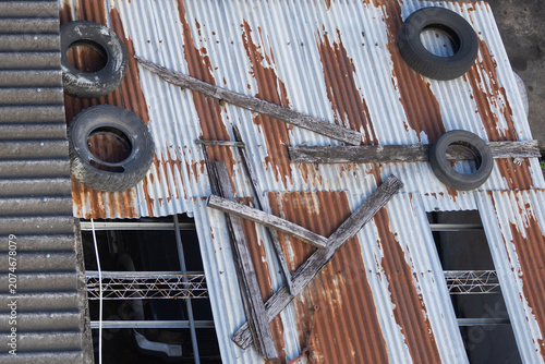 An old corrugated metal roof seen from above, with pieces of wood and tires on top. The corrugated sheets are discolored and old, with holes, the tires are worn out.