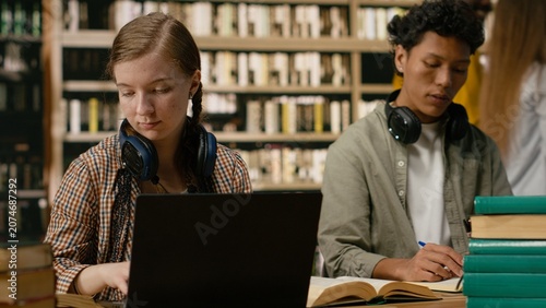 Εκτύπωση καμβά Young woman student reading and working on laptop between open books at library table with shelves behind her for digital learning and focused academic research for academic learning themes