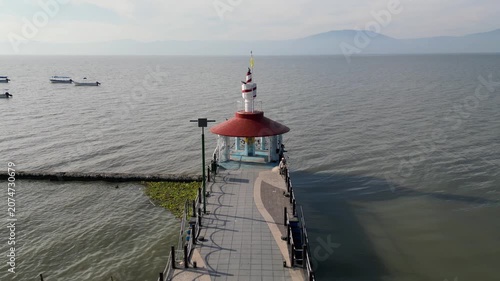 Drone view of Lake Chapala, Mexico. A pier with a red-roofed lighthouse. Boats on water. Hazy mountains. Serene scene