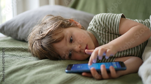 Little boy lying on bed and using smartphone for watching cartoons or playing games at home.