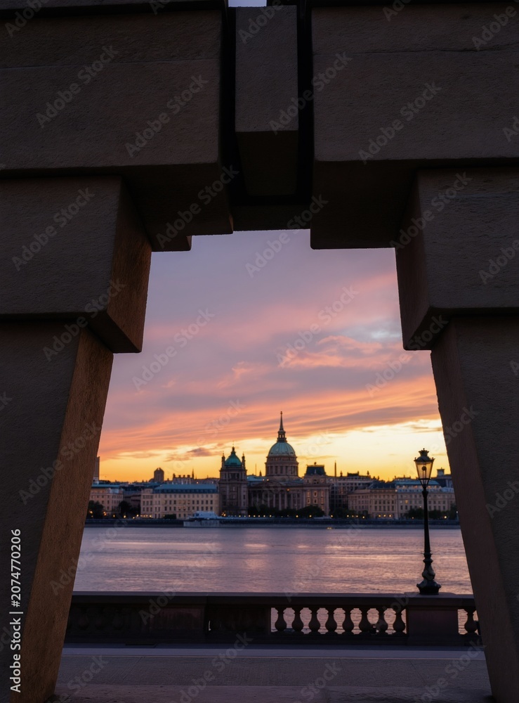 Fototapeta premium Stone monument frames historic cityscape across river at sunset. Pastel sky with clouds. Ornate buildings and classic lamppost visible. Calm water reflects twilight glow.
