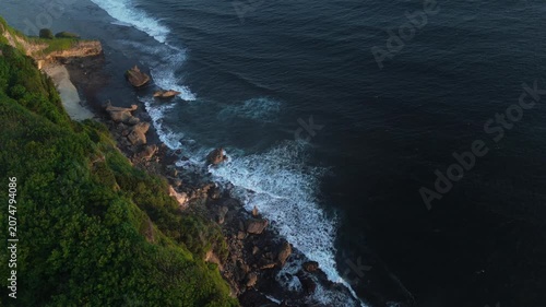 High angle aerial shot of dark sea waves splashing with white foam against green coastal rocks in Bali. Cinematic natural textures and contrast.