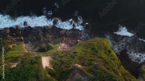 Spectacular top down drone view of powerful ocean waves crashing on rocky cliffs in Bali at sunset. Beautiful tropical shoreline scenery.