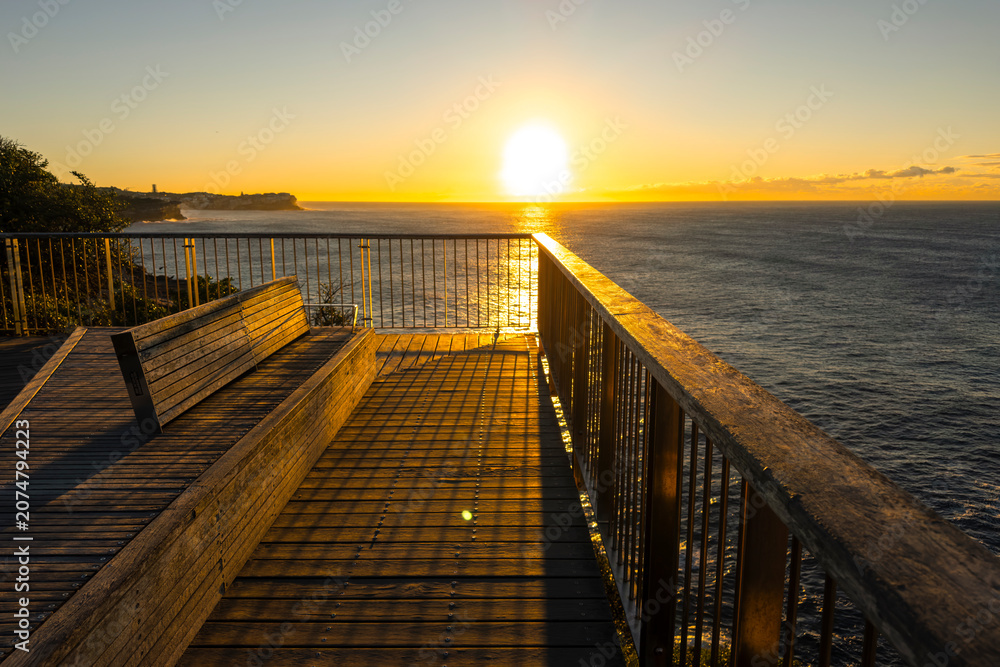 Obraz premium Sunrise over the Pacific Ocean viewed from the Bondi to Coogee Coastal Walk with golden light reflecting across calm water, Sydney, Australia.