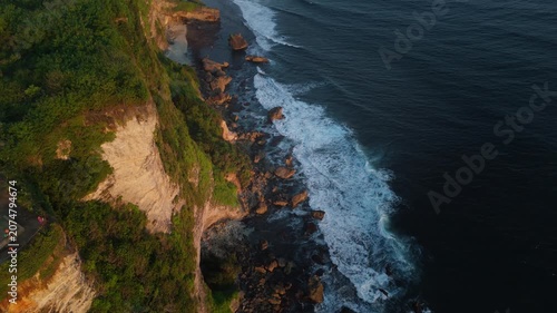 High angle drone shot of steep green cliffs meeting deep blue sea surf in Bali at dusk. Breathtaking tropical island coastline scenery.