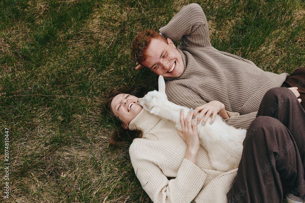Obraz premium Joyful couple lying on grass with a goat kid, enjoying an outdoor lifestyle, bonding and happiness in a relaxed moment, soft light, cozy sweaters, close connection, nature and playful calm.