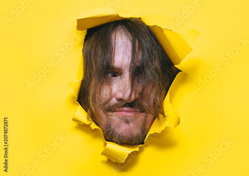 Foto A long-haired, shaggy man with a beard and mustache looks out from a torn hole in yellow paper with a strange expression on his face and a sly smile