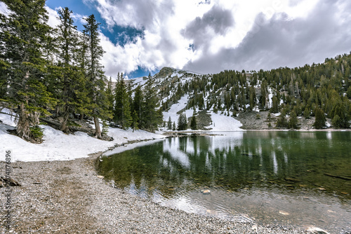 A serene wide-angle view of Teresa Lake surrounded by melting alpine snow and evergreen pine trees under a cloudy sky in Great Basin National Park.