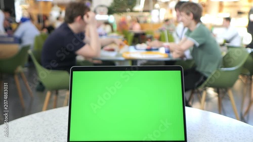 Laptop with green screen sits on cafe table. Background shows blurred people chatting and eating. Focus remains on laptop's screen. Useful for digital content, presentations, or mockups