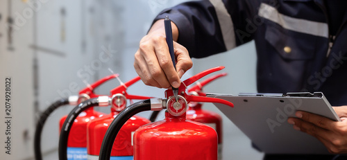 Technical engineer in uniform performing routine inspection on a row of red fire extinguishers during safety service check in industrial building.