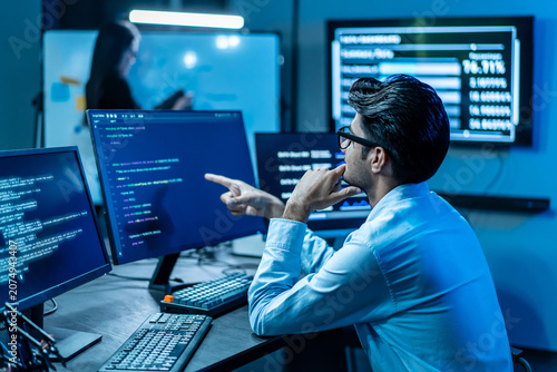 Photos Focused software developer analyzing code on multiple computer monitors in a dark modern tech office at night