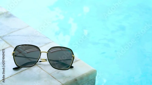 Luxury sunglasses resting on a marble pool tile against clear blue rippling water. Perfect summer holiday, travel, and luxury resort background for eyewear or lifestyle themes